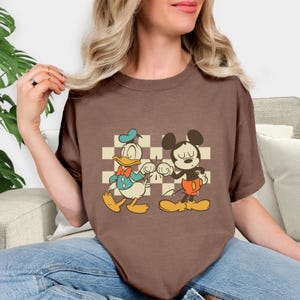 May include: A brown t-shirt featuring a retro cartoon graphic of Donald Duck and Mickey Mouse bumping fists. The characters are set against a checkered background. The shirt has short sleeves and a crew neck.