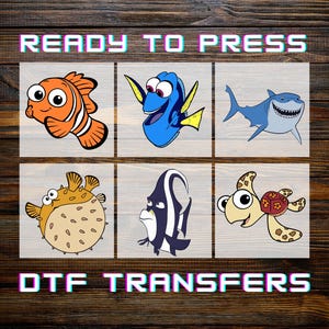 May include: DTF transfers featuring cartoon fish characters, including Nemo, Dory, and a shark, with the text "READY TO PRESS" and "DTF TRANSFERS" in a pixelated font. The image is set against a wood grain background.
