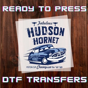 May include: A DTF transfer featuring a blue cartoon car with the text "Fabulous Hudson Hornet". The design includes the words "Piston Cup Champion '51 '52 '53". The words "READY TO PRESS" and "DTF TRANSFERS" are also visible.