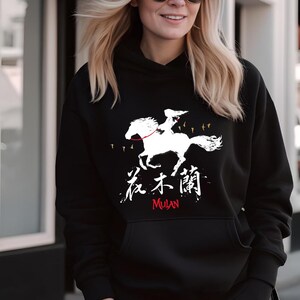 Pode incluir: Moletom preto com um gr&aacute;fico branco de uma pessoa montando um cavalo, caracteres chineses e a palavra "Mulan" em vermelho. O design inclui p&aacute;ssaros em voo. Um moletom confort&aacute;vel e cl&aacute;ssico.