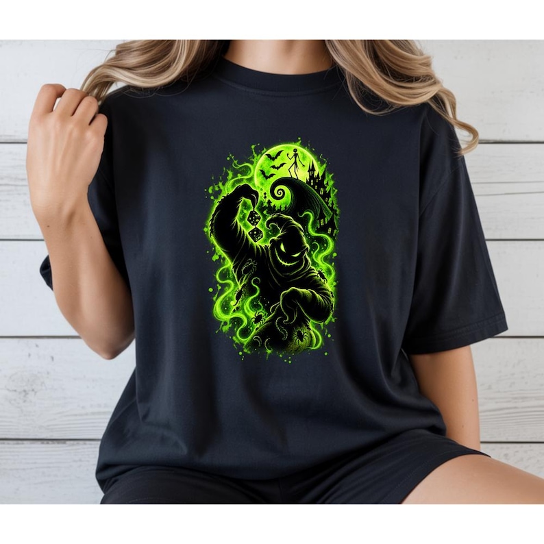 May include: A black t-shirt featuring a vibrant green graphic print of a sack-like character holding dice. The design includes a glowing moon, bats, a crooked house silhouette, and swirling smoke effects surrounding the central figure.