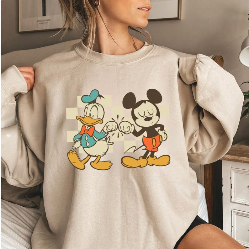 May include: Beige sweatshirt featuring a graphic of Donald Duck and Mickey Mouse bumping fists. Donald Duck is in a blue sailor suit with a red bow tie. Mickey Mouse wears red shorts and yellow shoes. The cartoon characters are set against a checkered background.