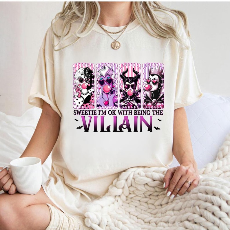May include: Cream-colored t-shirt featuring a graphic of four cartoon villains blowing bubblegum bubbles. The text reads "Sweetie I'm OK with being the Villain" in purple and black. The shirt is a casual, short-sleeved style.