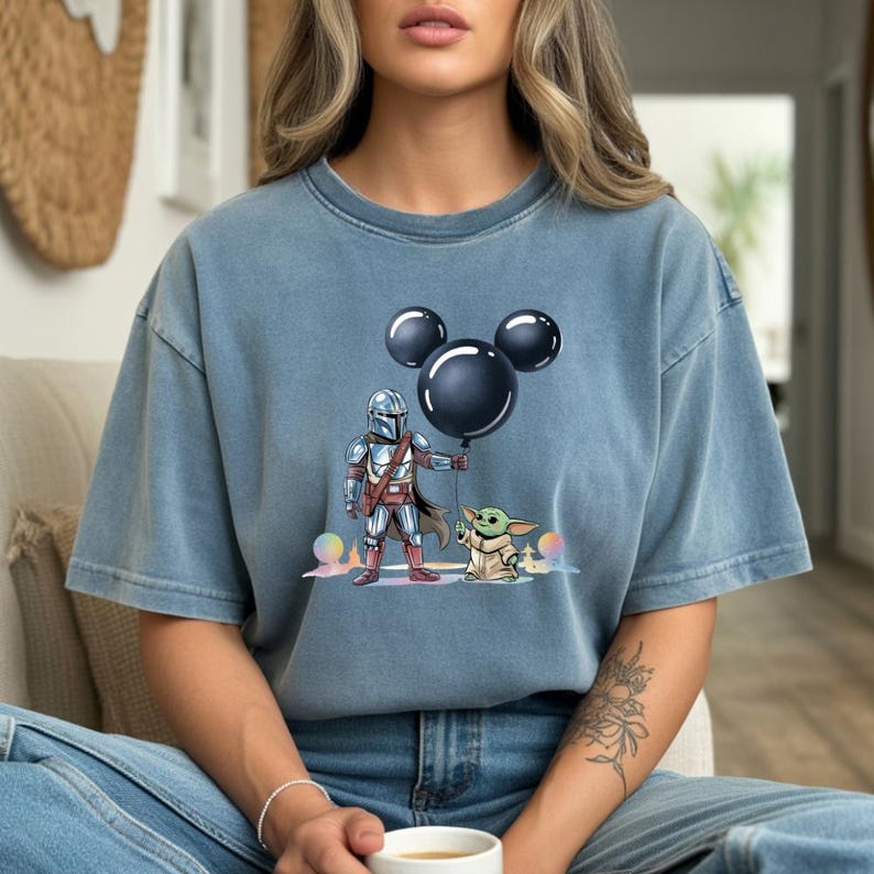 May include: A blue t-shirt features a graphic of The Mandalorian holding a black balloon shaped like Mickey Mouse ears, with Grogu standing beside him. The design is in a cartoon style. The shirt is a solid blue color.