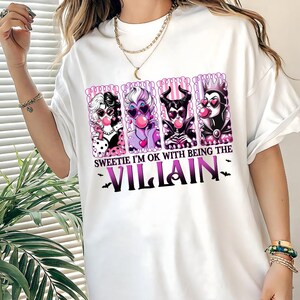 May include: White t-shirt featuring a graphic of four cartoon villains blowing bubblegum bubbles. The design includes the text "Sweetie I'm OK with being the Villain" in purple and black, with bat silhouettes.