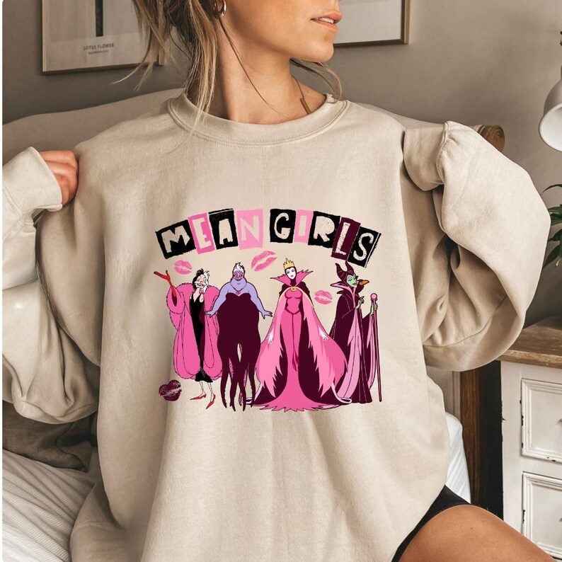 May include: Beige sweatshirt with the text "MEAN GIRLS" in black and pink block letters. The graphic features illustrations of four cartoon villains with pink and purple details, and red lipstick kisses.