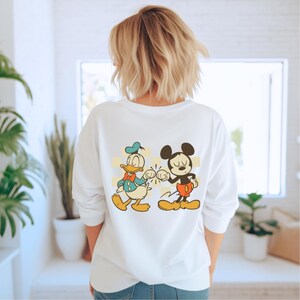 May include: White long-sleeve sweatshirt featuring a graphic of Donald Duck and Mickey Mouse bumping fists. Donald Duck is wearing a blue hat and bow tie. Mickey Mouse is wearing red shorts and yellow shoes. The design is set against a yellow and white checkered background.
