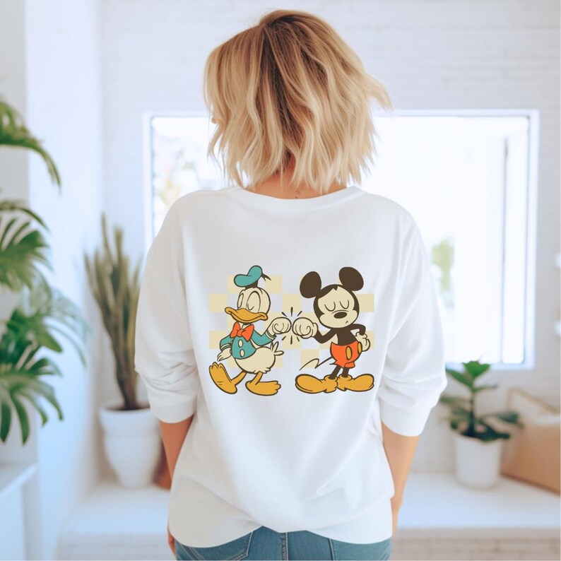 May include: White long-sleeve sweatshirt featuring a graphic of Donald Duck and Mickey Mouse bumping fists. Donald Duck is wearing a blue hat and bow tie. Mickey Mouse is wearing red shorts and yellow shoes. The design is set against a yellow and white checkered background.