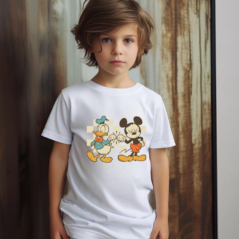 May include: A white t-shirt featuring a cartoon graphic of Mickey Mouse and Donald Duck bumping fists. The characters are set against a yellow and white checkered background. The shirt is being worn by a child.