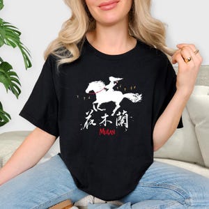 Pode incluir: T-shirt preta com um gr&aacute;fico branco que retrata uma pessoa montada a cavalo, caracteres chineses e a palavra "MULAN" em vermelho. O design inclui pequenas silhuetas de p&aacute;ssaros. Uma camiseta casual e confort&aacute;vel.