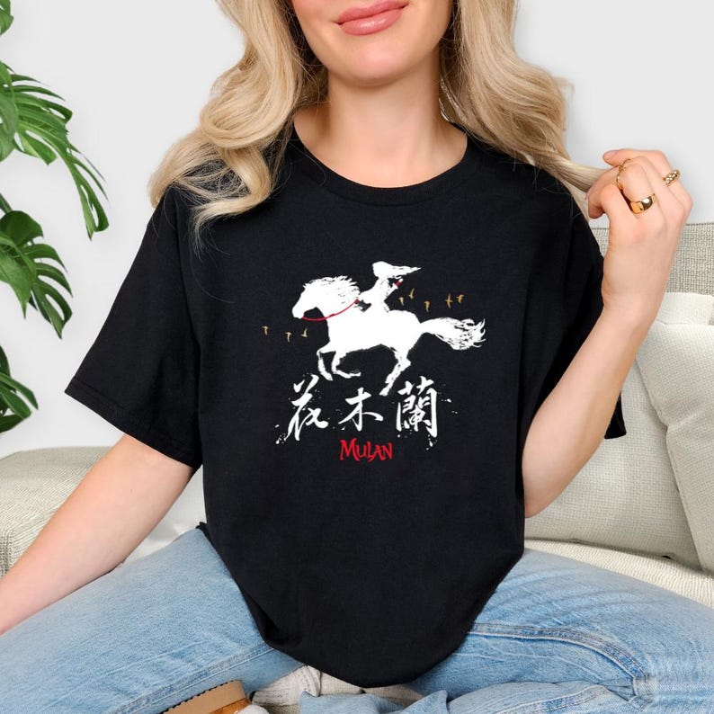 Pode incluir: T-shirt preta com um gr&aacute;fico branco que retrata uma pessoa montada a cavalo, caracteres chineses e a palavra "MULAN" em vermelho. O design inclui pequenas silhuetas de p&aacute;ssaros. Uma camiseta casual e confort&aacute;vel.