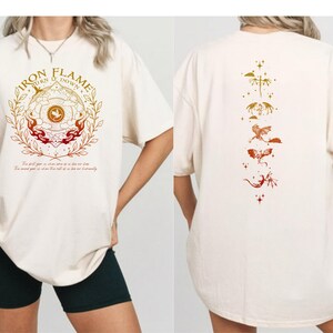 Op de afbeelding: Cr&egrave;mekleurig T-shirt met een grafische print op de voorkant met de tekst "IRON FLAME" en een cirkelvormig ontwerp met vlammen en botanische elementen. De achterkant van het shirt toont een verticale opstelling van gestileerde draken en sterren in oranje en goud.