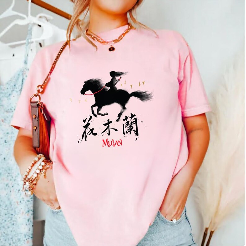 Pode incluir: T-shirt rosa claro com uma silhueta preta de uma pessoa montando um cavalo, com o texto "花木蘭" e "MULAN" em vermelho. O design &eacute; uma representa&ccedil;&atilde;o gr&aacute;fica da personagem da Disney Mulan.