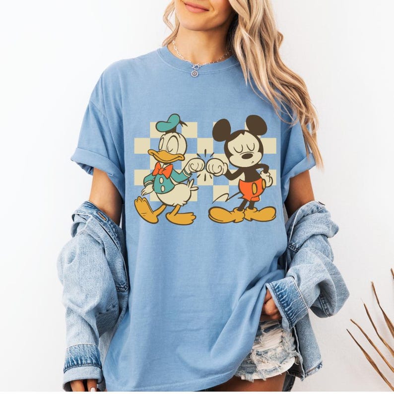 May include: Light blue t-shirt featuring a retro graphic of Donald Duck and Mickey Mouse bumping fists. The cartoon characters are set against a checkered background. The shirt has rolled-up sleeves and a relaxed fit.