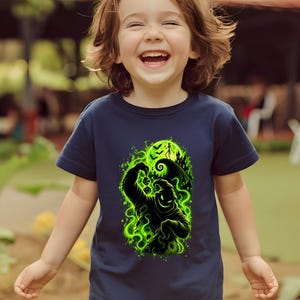 May include: A navy blue short-sleeved t-shirt featuring a vibrant green graphic print of a shadowy, sack-like character holding dice, surrounded by swirling smoke, spiders, a crescent moon, flying bats, and a silhouette of a crooked house.
