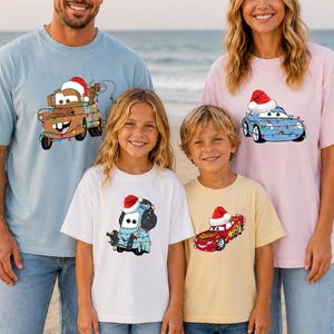 A family of four, consisting of two adults and two children, are standing on a beach wearing matching t-shirts with cartoon characters from the Cars movie.