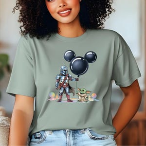 May include: Sage green t-shirt featuring a cartoon illustration of The Mandalorian and Grogu. The Mandalorian is holding a black Mickey Mouse balloon, and Grogu is standing next to him. The design includes colorful orbs and a pastel background.