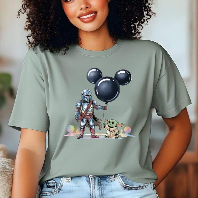 May include: Sage green t-shirt featuring a cartoon illustration of The Mandalorian and Grogu. The Mandalorian is holding a black Mickey Mouse balloon, and Grogu is standing next to him. The design includes colorful orbs and a pastel background.