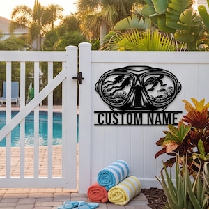 May include: A black metal wall sign shaped like a diving mask, featuring cut-out silhouettes of a scuba diver and a sea turtle within the lenses. Below the mask, a horizontal bar displays the text CUSTOM NAME in bold, capital letters.