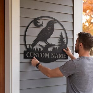 May include: A circular black metal wall sign featuring a silhouette of a raven perched on a branch, a crescent moon, and pine trees. Below the circular frame is a rectangular plaque with the text CUSTOM NAME in bold, capital letters.