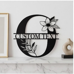 Puede incluir: Monograma de metal negro con la letra "O" con detalles florales y de hojas. La letra presenta el texto "CUSTOM TEXT" en el centro. Una flor decorativa y hojas están en el lado derecho e inferior izquierdo. El monograma se muestra sobre una superficie blanca.