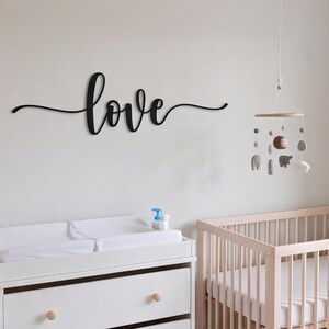 Puede incluir: Un letrero de pared negro con la palabra love en una fuente cursiva. Al lado cuelga un móvil con un marco circular de madera y formas de fieltro colgantes, que incluyen un planeta marrón, animales grises y nubes blancas.