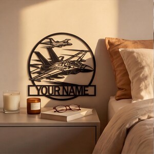 May include: A black circular metal wall art piece featuring two fighter jets in flight, with a rectangular nameplate at the bottom containing the text "YOUR NAME" in bold, capital letters. The design is cut from a single sheet of metal.