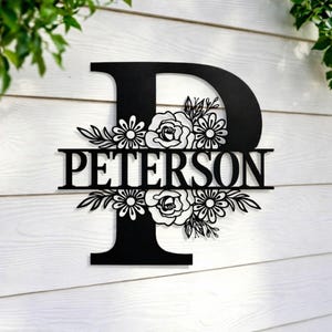 May include: Black metal monogram letter "P" with the name "PETERSON" cut out. The letter is decorated with floral designs, including roses and daisies. The sign is mounted on a white wooden background.