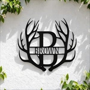 May include: Black metal wall art featuring the name "BROWN" within a letter "D" and framed by stylized antlers. The artwork is mounted on a white textured wall, with greenery in the corners.