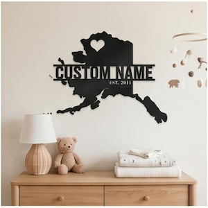 May include: A black metal wall art piece shaped like the map of Alaska, featuring a heart cutout, a horizontal bar with the text CUSTOM NAME, and the text EST. 2011. The design is a flat, silhouette-style cutout made of dark, solid material.