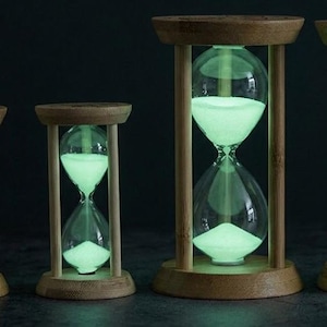 May include: Four hourglasses with wooden frames and clear glass bulbs containing glowing light green sand. The two smaller hourglasses are on the left, and the two larger hourglasses are on the right, all arranged in a row against a dark background.