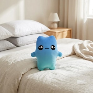 May include: A soft plush toy shaped like a stylized creature with a gradient blue color transitioning from dark to light. The toy features large black embroidered eyes with white highlights and a simple curved line for a mouth, with a small white tag.