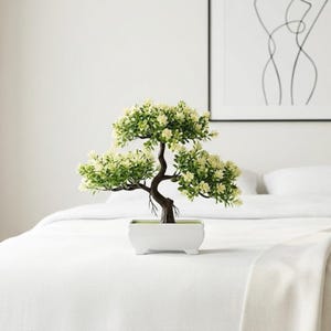 May include: A small artificial bonsai tree featuring a dark brown, curved trunk and lush green foliage with tiny cream-colored flowers. The plant is potted in a rectangular, white ceramic container with four small feet, creating a balanced display.