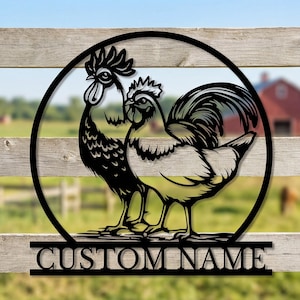 May include: Black metal rooster silhouette art piece. Two roosters are inside a circular frame, with the text "CUSTOM NAME" below. The artwork is mounted on a wooden fence, with a blurred background of a field and a red barn.