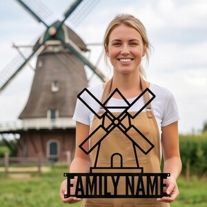 May include: A black metal windmill sign with the text "FAMILY NAME" is held by a person. The sign features a windmill design with blades and a base. A windmill is in the background, along with green grass and a cloudy sky.