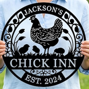 Puede incluir: Letrero de metal negro con un diseño de gallina y polluelos, el texto "JACKSON'S CHICK INN EST. 2024" se muestra en un marco circular. El letrero se sostiene frente a un fondo verde borroso.