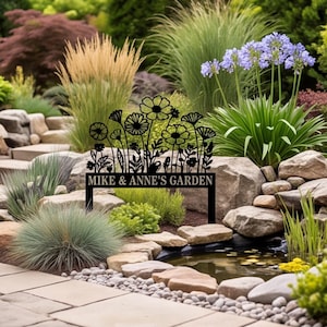 Op de afbeelding: Een zwart metalen tuinbord met een silhouet van diverse wilde bloemen en stengels. De rechthoekige basis van het bord toont de tekst "MIKE & ANNE'S GARDEN" in een duidelijk hoofdlettertype, ondersteund door twee verticale pinnen voor in de grond.