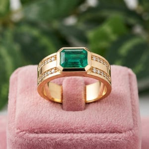 Peut inclure: Une bague en or ornée d'une pierre précieuse verte taille émeraude sertie dans une monture. L'anneau est agrémenté de deux rangées de petites pierres rondes et transparentes incrustées le long de la tige en or, offrant un aspect poli.