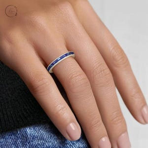 May include: A silver-toned band ring featuring a continuous row of small, rectangular-cut blue gemstones set in a channel setting. The ring is worn on a finger, showcasing the uniform alignment of the vibrant blue stones along the metallic band.