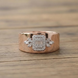 Op de afbeelding: Een brede ring van roségoud met een centraal rechthoekig cluster van kleine ronde en baguette-geslepen diamanten. De diamantzetting is uitgevoerd in wit metaal, wat een contrast vormt met het gepolijste roségouden oppervlak van de ring.
