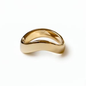 Puede incluir: Un anillo de oro pulido con una superficie lisa y continua, que presenta un diseño de banda ondulado y sinuoso. El metal tiene un acabado brillante y reflectante que capta la luz, creando reflejos suaves a lo largo de la forma orgánica.