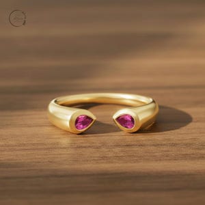 Könnte beinhalten: Ein offener Goldring mit zwei tropfenförmigen rosa Edelsteinen an den Enden der Ringschiene. Der Ring hat eine glatte, polierte Oberfläche und ein abgerundetes Profil, wobei die leuchtend rosa Steine einen Kontrast zum Gold bilden.