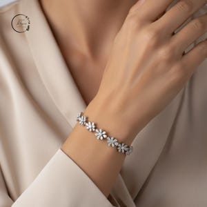 May include: A silver-toned bracelet featuring a series of connected flower-shaped links, each encrusted with multiple clear, sparkling stones. The design consists of repeating floral motifs arranged in a continuous line around the wrist.