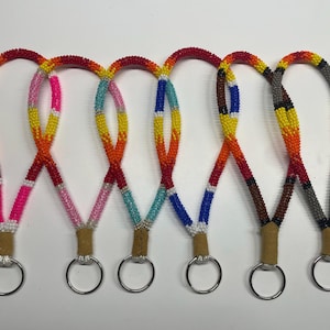 May include: Six wristlet keychains made of small, colorful glass beads arranged in various patterns, including gradients of pink, orange, yellow, blue, and brown. Each loop is secured with a tan leather tab and a circular silver metal key ring.