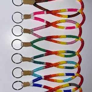 May include: Seven beaded keychains in various colors, including brown, pink, green, blue, and gray. Each keychain features a silver ring and a leather loop. The beads are arranged in colorful patterns, creating a vibrant and eye-catching design.