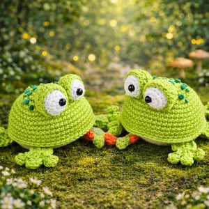 Puede incluir: Dos ranas de ganchillo hechas a mano, de color verde lima, con ojos blancos y detalles en naranja. Están colocadas sobre un lecho de musgo verde y flores blancas, con un fondo borroso de árboles y sol.