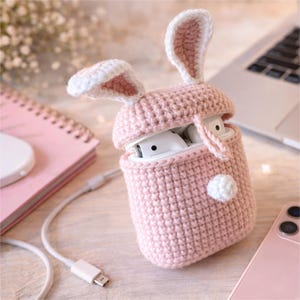 May include: A pink crocheted bunny ear case for wireless earbuds. The case is open, revealing white earbuds inside. The bunny ears are white with pink interiors, and a small white pom-pom is attached to the side. A charging cable and a pink phone are also visible.