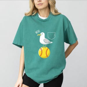 Può includere: T-shirt verde acqua con una grafica di un gabbiano su una palla da softball gialla. La parola "MINE" è sopra il gabbiano. Un contorno di tasca bianco è a destra del gabbiano.