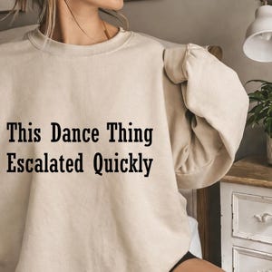 Może przedstawiać: Beżowa bluza z napisem "This Dance Thing Escalated Quickly" czarnymi literami. Bluza ma neutralny kolor i długie rękawy. Zdjęcie przedstawia osobę noszącą bluzę.