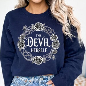 Puede incluir: Sudadera azul marino con un diseño floral y de espinas. El texto "THE DEVIL HERSELF" está centrado. El diseño presenta rosas y espinas.